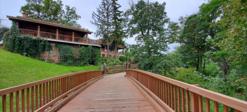 A wooden bridge leads to a lush green area with trees and a rustic building in the background.