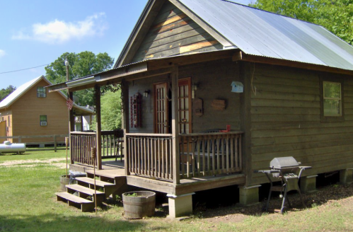A rustic wooden cabin with a porch, steps, and a grill, surrounded by greenery and blue skies.