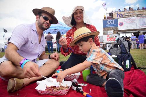 A family enjoys a meal together outdoors, sitting on a blanket with food and wearing hats at a festive event.