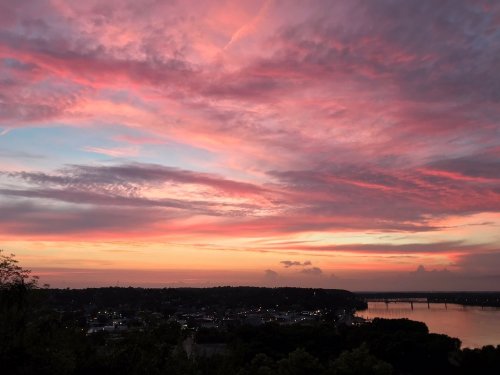 A vibrant sunset with pink and orange clouds over a river and a small town, creating a serene evening atmosphere.