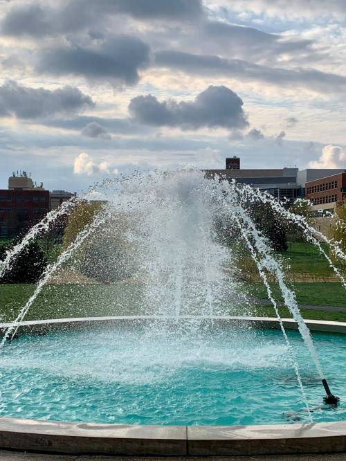 A fountain sprays water into a turquoise pool, with a cloudy sky and buildings in the background.
