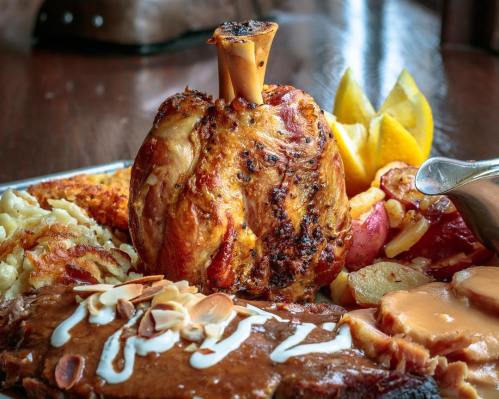 A platter featuring a roasted meat dish with a bone, surrounded by sides like potatoes, apples, and lemon wedges.