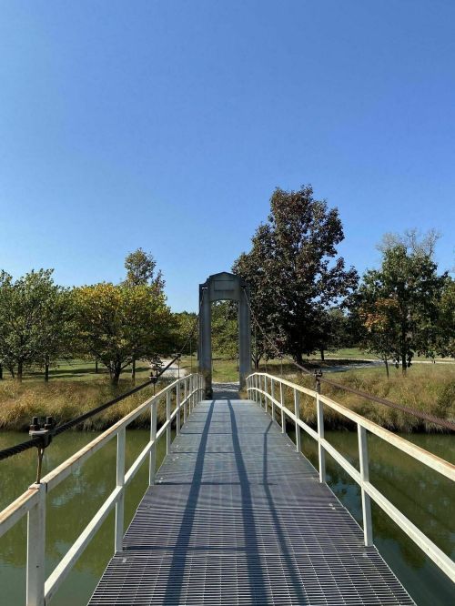 A suspension bridge over a calm waterway, surrounded by trees under a clear blue sky.