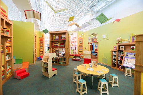 Bright children's library with colorful bookshelves, playful seating, and a round table surrounded by small stools.