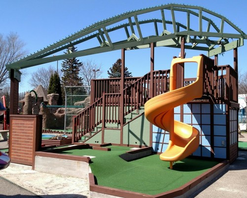 Colorful playground structure featuring a spiral slide, climbing areas, and a green roof against a clear blue sky.