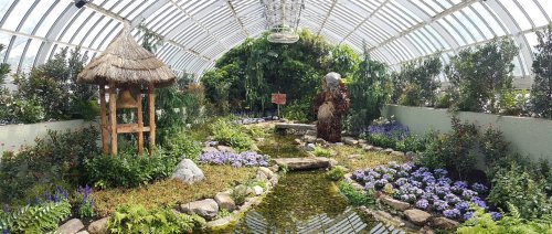 Lush indoor garden with a pond, colorful flowers, a thatched gazebo, and a stone statue surrounded by greenery.