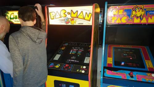 A person stands at a classic Pac-Man arcade machine, with Ms. Pac-Man and another arcade game visible nearby.