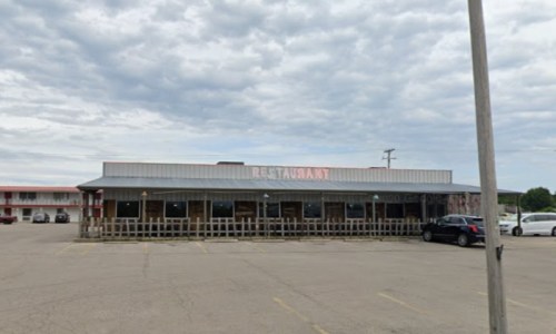 A restaurant building with a metal roof, large windows, and a parking lot, under a cloudy sky.