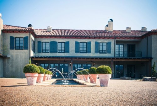 A modern building with a red-tiled roof, blue shutters, and a fountain surrounded by potted plants in a gravel courtyard.