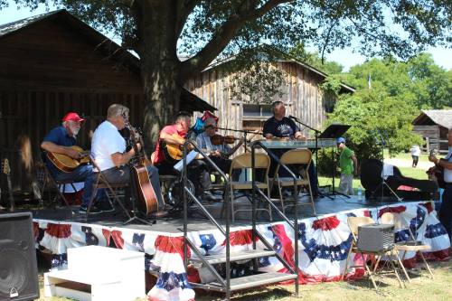 A band performs on a stage decorated with red, white, and blue, surrounded by rustic buildings and an audience.