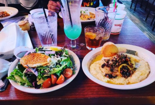 A table with a salad, a plate of grits topped with shrimp, and colorful drinks in glasses.