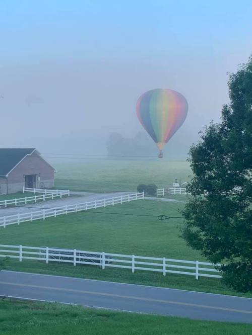 A colorful hot air balloon floats above a misty field, with a farmhouse and white fences in the foreground.
