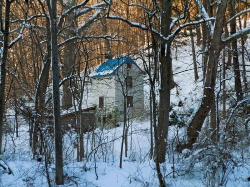A snow-covered house nestled among trees in a winter landscape, with a blue roof and warm sunlight filtering through.