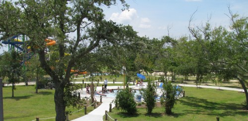 A sunny park scene with water slides, a pool area, and people enjoying the outdoors among trees and green grass.