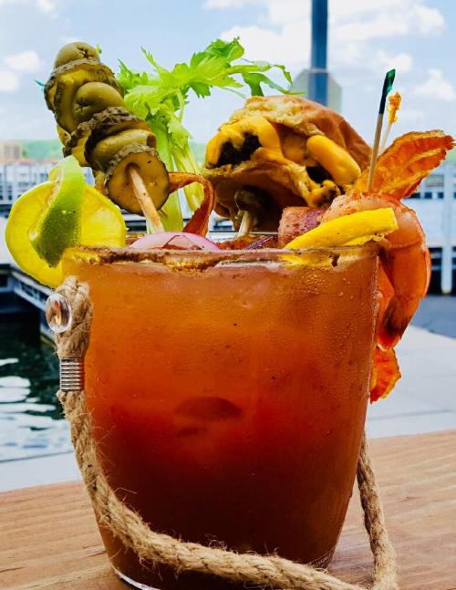 A vibrant Bloody Mary garnished with shrimp, pickles, lemon, and a mini burger, served in a rustic glass by the water.