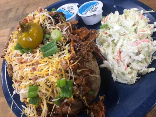 A loaded baked potato topped with pulled pork, cheese, green onions, and a jalapeño, served with coleslaw and sauces.