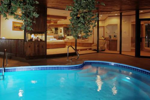 Indoor pool with a blue water, surrounded by plants, leading to a cozy room with a fireplace and large windows.