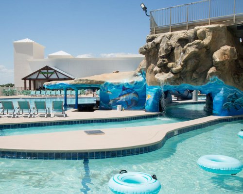 A sunny pool area featuring a rock formation, water slide, and floating tubes, surrounded by lounge chairs.