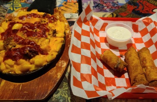 A sizzling plate of cheesy mac and cheese topped with BBQ sauce, alongside crispy egg rolls and a side of ranch dressing.