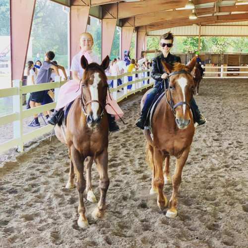 Two riders on horseback in an indoor arena, smiling and enjoying their time, with people in the background.