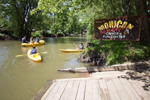 A group of people kayaking on a river near a sign for Mohican Adventures Canoe & Fun Center. Lush greenery surrounds the area.