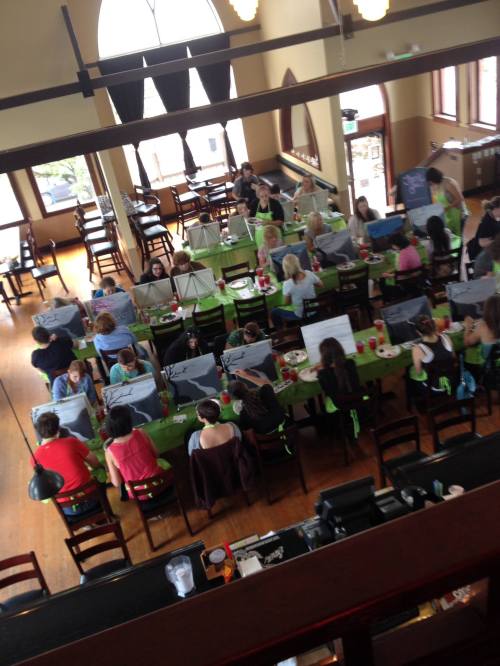 Aerial view of a lively painting class in a café, with participants at tables, canvases, and drinks.