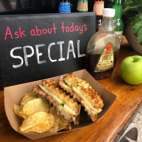 A sandwich with apple and cheese, chips, a green apple, and maple syrup, next to a sign reading "Ask about today's SPECIAL."