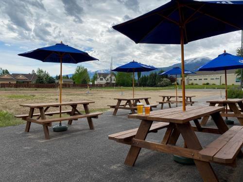 Outdoor seating area with wooden picnic tables and blue umbrellas, overlooking a grassy field and mountains in the background.
