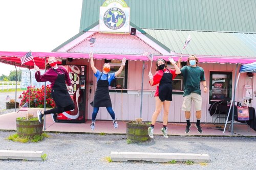 Four people in masks jump in front of a colorful restaurant, holding small American flags, celebrating together.