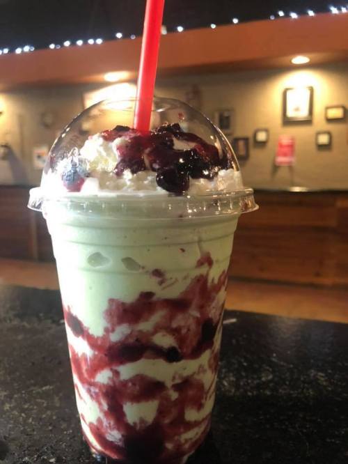 A creamy green milkshake topped with whipped cream and berry sauce, served in a clear cup with a red straw.