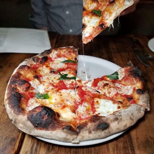 A freshly baked pizza with melted cheese, tomato sauce, and basil, with a slice being lifted, on a wooden table.