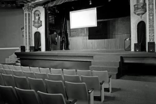 A dimly lit theater stage with a blank screen and empty seats in the foreground.