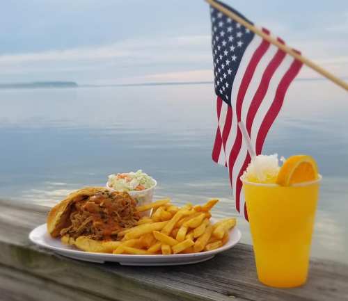 A plate of pulled pork sandwich, fries, and coleslaw, with a drink and a small American flag by a calm waterfront.