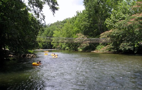 A serene river scene with people floating on yellow tubes, surrounded by lush green trees and a hanging bridge above.