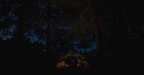 A brightly lit tent in a dark forest, surrounded by tall trees under a night sky.