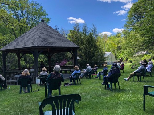 A group of people sitting in chairs outdoors, listening to a speaker under a gazebo surrounded by greenery.