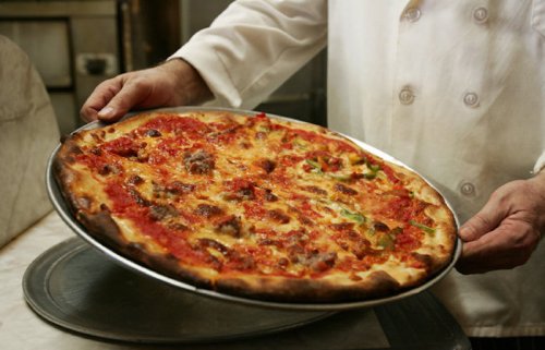 A chef holds a freshly baked pizza on a metal tray, showcasing its melted cheese and toppings.