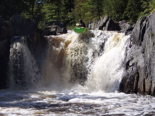 A kayaker descends a waterfall, surrounded by rocky cliffs and rushing water in a lush, green forest.