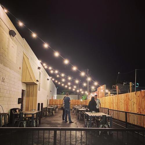 Outdoor dining area at night, featuring string lights, empty tables, and two people conversing near a wooden fence.