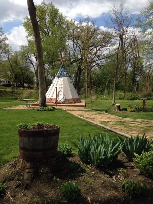 A teepee stands in a green park surrounded by trees, with a garden and a path leading to it.