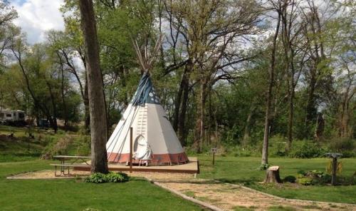 A traditional teepee stands in a green park surrounded by trees, with a picnic table nearby.