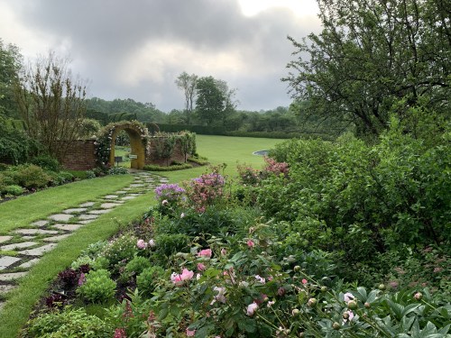 A lush garden path leads through vibrant flowers and greenery, with a cloudy sky in the background.