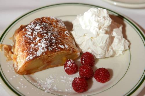 A slice of pastry dusted with powdered sugar, served with whipped cream and fresh raspberries on a plate.