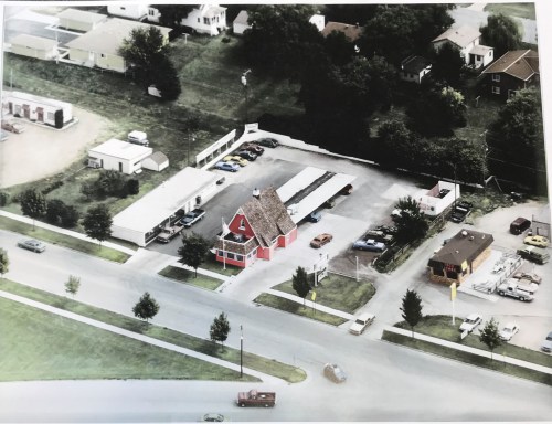 Aerial view of a red building with a peaked roof, surrounded by parking and greenery, near a road intersection.