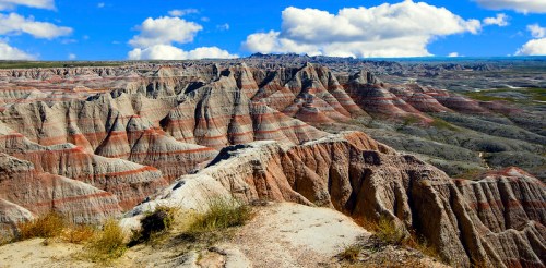 A panoramic view of colorful, layered rock formations under a bright blue sky with fluffy clouds.