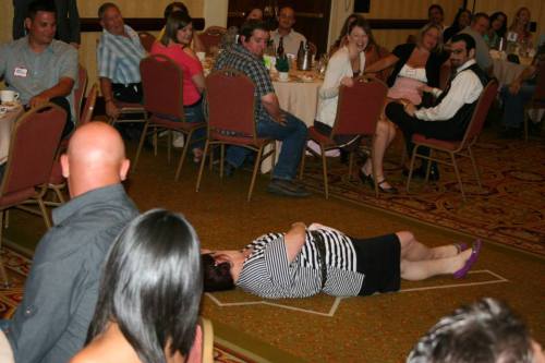 A woman in a striped dress lies on the floor while a group of people at tables watches and laughs.