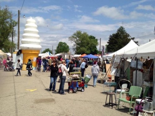 A bustling outdoor market with tents, people browsing, and a large ice cream cone statue in the background.