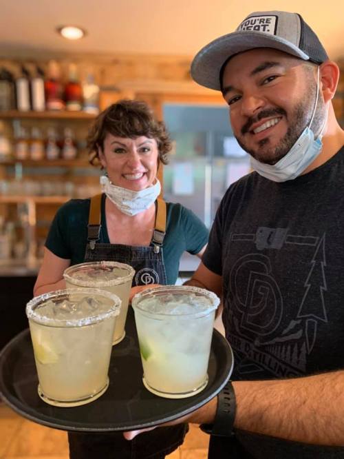 Two smiling bartenders hold a tray of margaritas in a cozy bar setting.