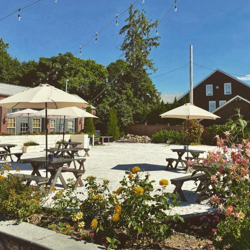 A sunny outdoor space with picnic tables, umbrellas, and blooming flowers, surrounded by greenery and string lights.