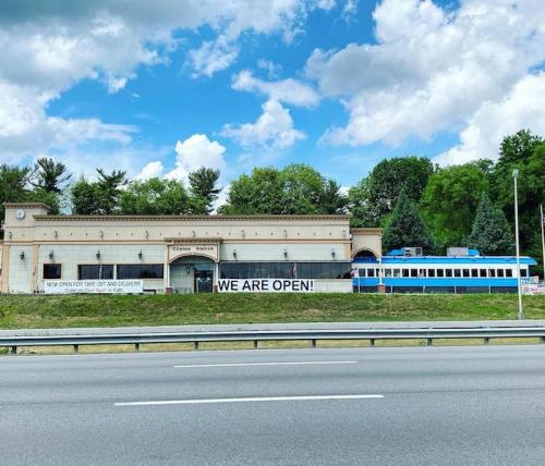 A building with a sign reading "WE ARE OPEN!" and a blue train parked beside it, set against a cloudy sky.